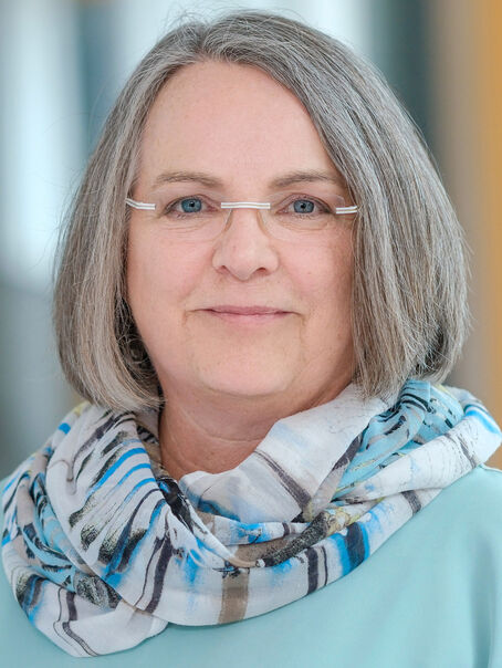 Das Bild zeigt eine weiblich lesbare Person mit kinnlangen grauen Haaren, einer Brille ohne Fassung, einem hellgemusterten Schal und einem hellblauen Pullover.