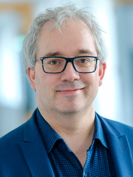 Eine männlich gelesene Person mit kurzen grauen Haaren und dunkler Brille. Er trägt ein dunkelblaues Hemd und ein blaues Sakko. 
